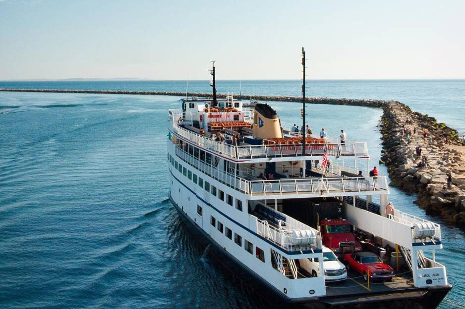 The Block Island Ferry, carrying cars and passengers, sails through a narrow channel between rocky breakwaters. The sky is clear and the sea, calm. This white multi-deck vessel has people visibly enjoying the stunning view.