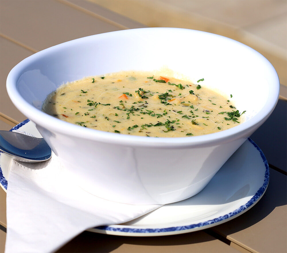 A white bowl of creamy soup, inspired by the Ballards Block Island Menu, is garnished with chopped herbs and sits on a white plate with a spoon and napkin, placed on a light brown table.