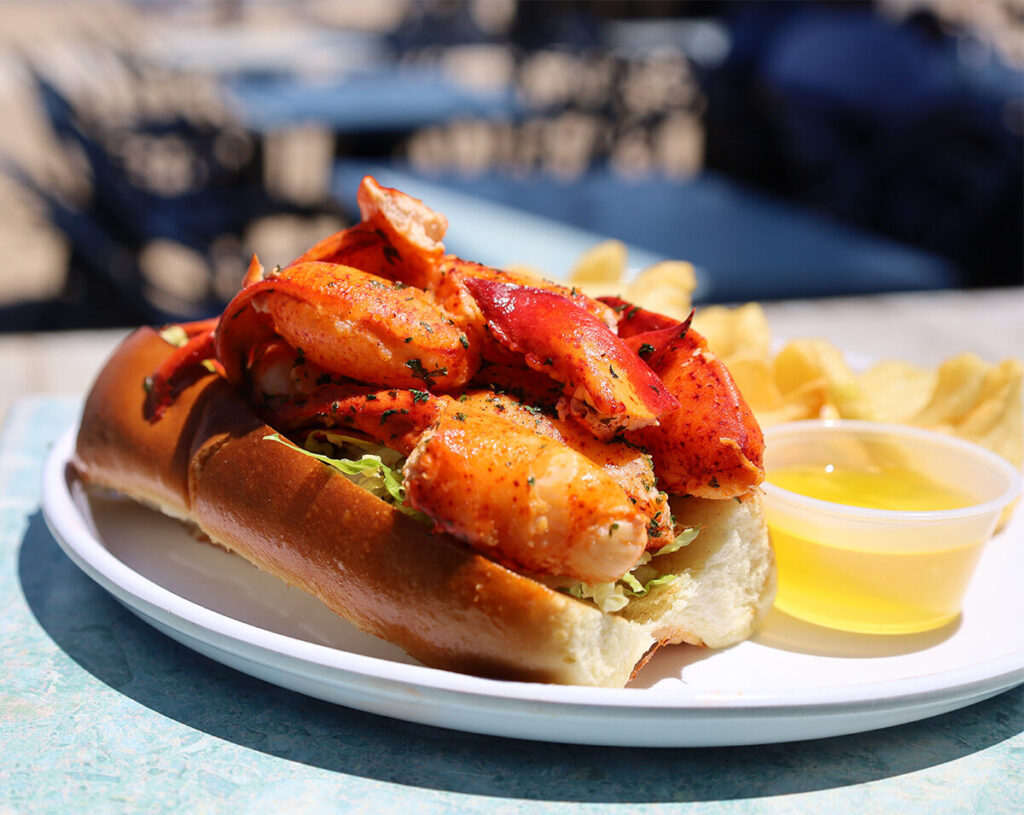 A lobster roll filled with lobster meat on a toasted bun, served with potato chips and melted butter on a white plate—just one of the fresh seafood favorites from the Ballards Block Island Menu.