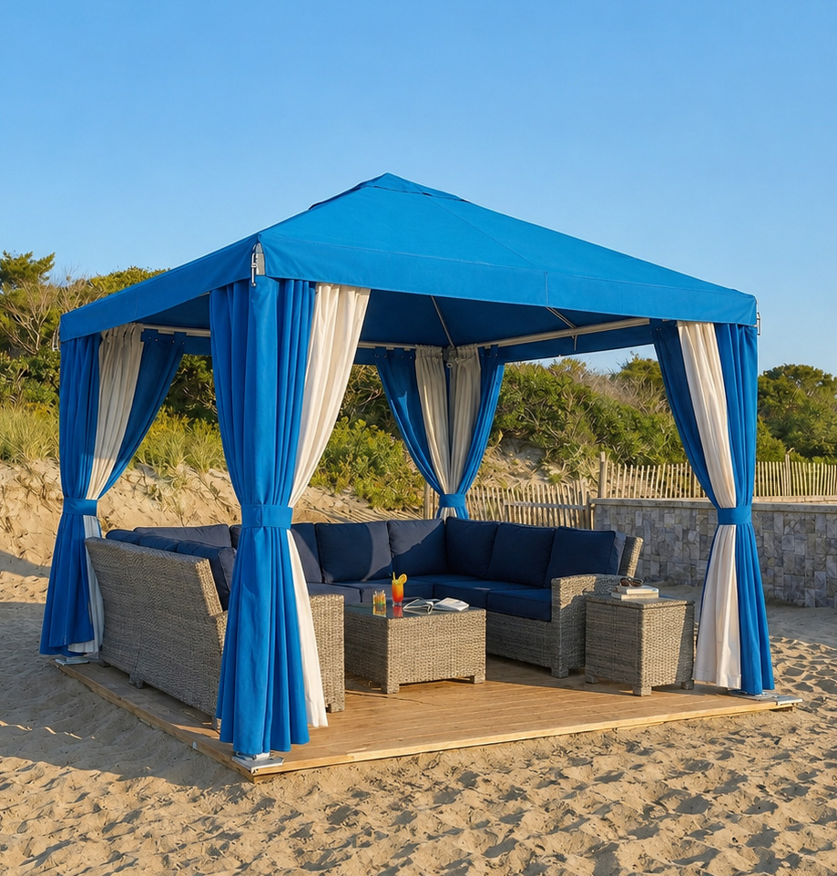 A blue and white outdoor canopy with curtains stands on a sandy beach at Block Island Beach Resort, covering wicker patio furniture with blue cushions. A tray with drinks rests on the coffee table under the bright, clear sky.