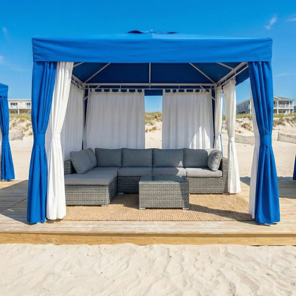 A blue and white cabana with gray outdoor furniture sits on a wooden platform on a sandy beach, with dunes and buildings visible in the background under a clear blue sky.